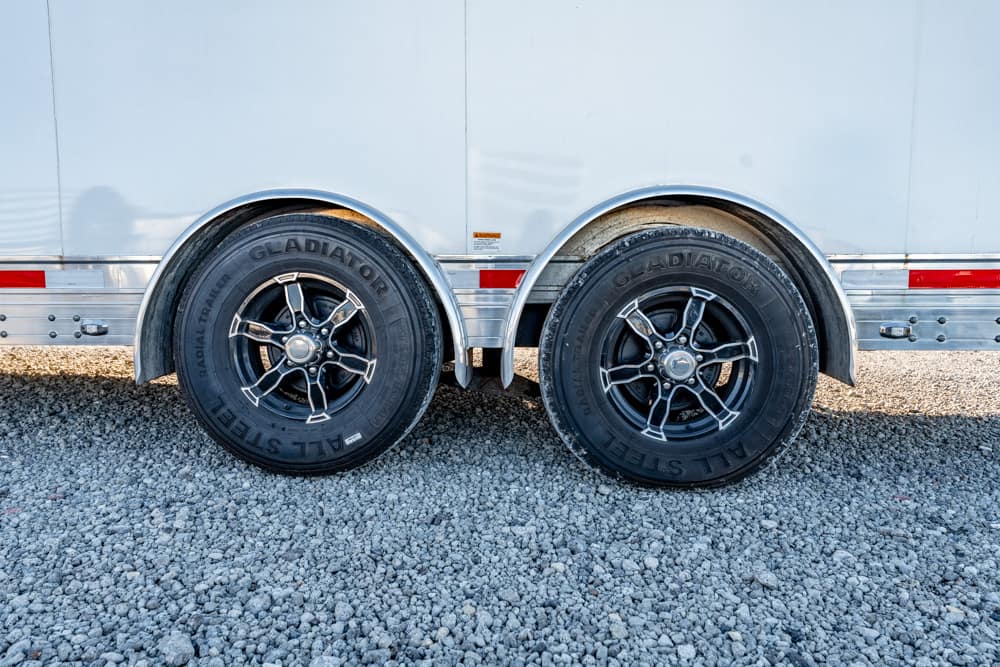 Dual-axle trailer with two black Gladiator wheels mounted on gravel, showing wheel wells and fenders in a close-up view.
