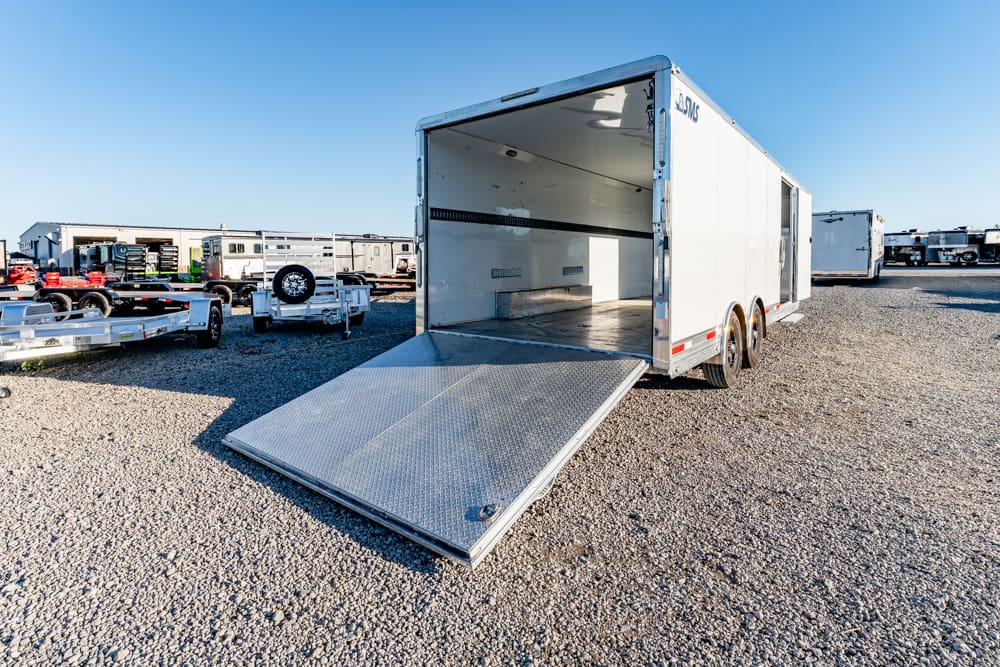 White enclosed cargo trailer with a metal ramp lowered at the rear, on a gravel lot under a clear blue sky.