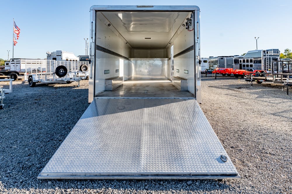 Open aluminum trailer with the diamond-plate ramp lowered to the ground, empty cargo area, in a gravel lot with other trailers and an American flag in the background.