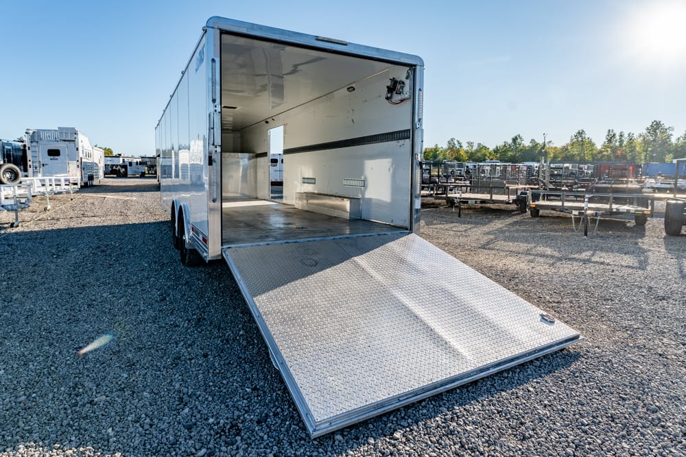 Open empty aluminum cargo trailer with ramp down on gravel lot, surrounded by other trailers in the background.