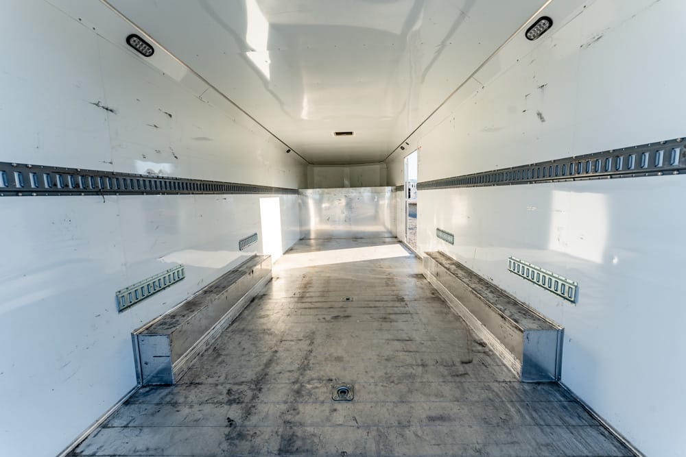 Interior of an empty white cargo trailer with metal side rails and a worn metal floor, viewed from the rear/doorway.