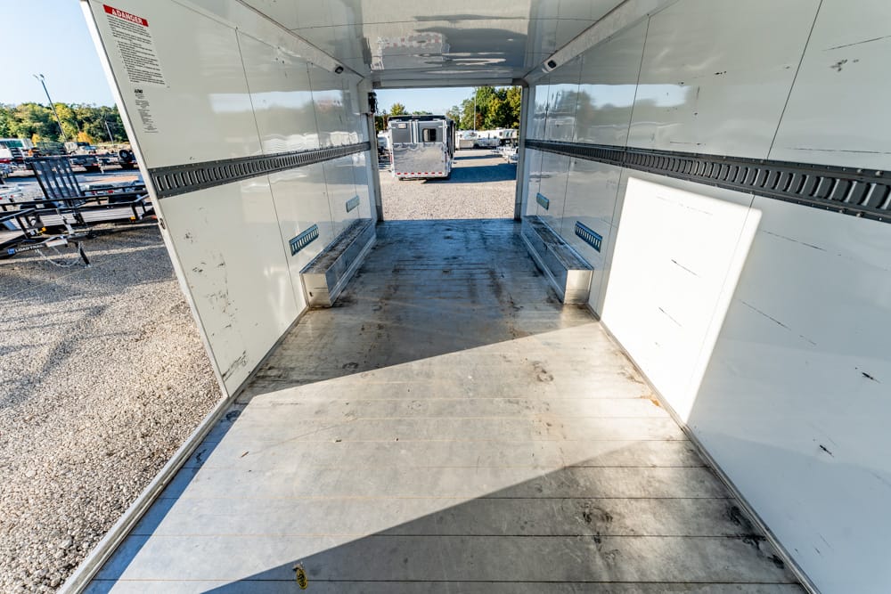 Interior of an empty cargo trailer with metal floor and white walls, looking toward the open back where other trailers are parked in a lot