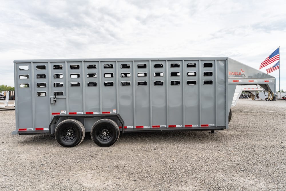 Side view of a large gray livestock trailer with ventilation slots, parked on a gravel lot with an American flag in the background accuracy: cattle trailer