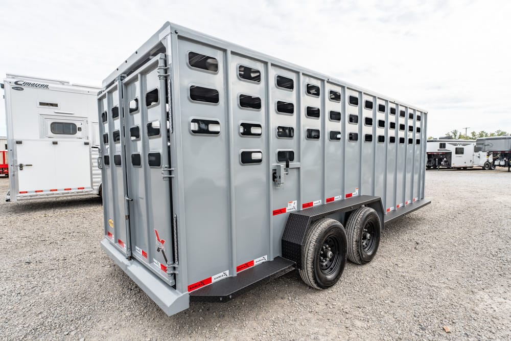 Gray livestock trailer with vertical vented panels and dual axles parked on a gravel lot, other trailers visible in the background