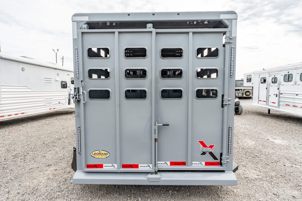 Rear of a gray livestock trailer with vented doors and small windows in a gravel lot, showing branding and latches
