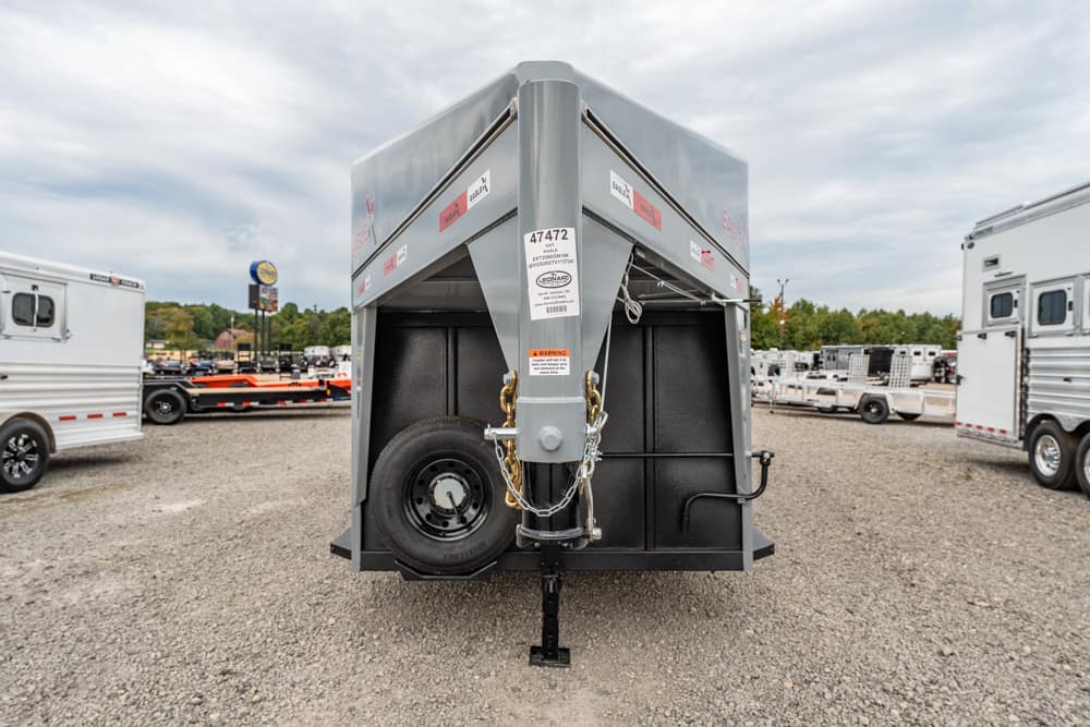 Rear view of a gray dump trailer with a spare tire, safety chains, and hitch on a gravel lot; other trailers visible in the background.