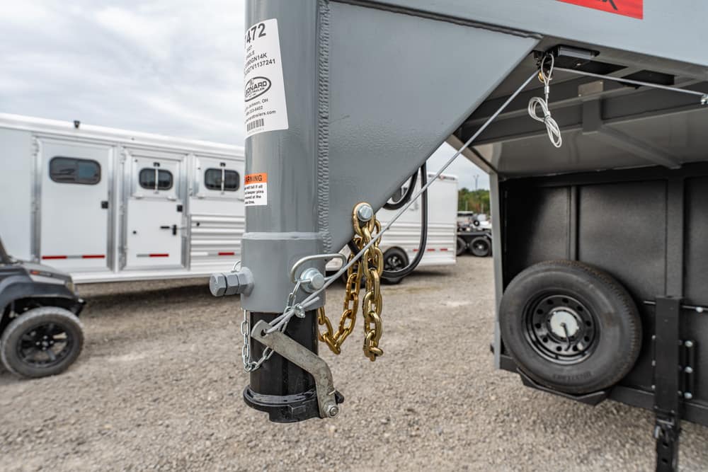 Close-up of a trailer hitch on a gooseneck trailer with safety chains, latch, and a coiled cable, spare tire mounted on the side in a gravel lot.
