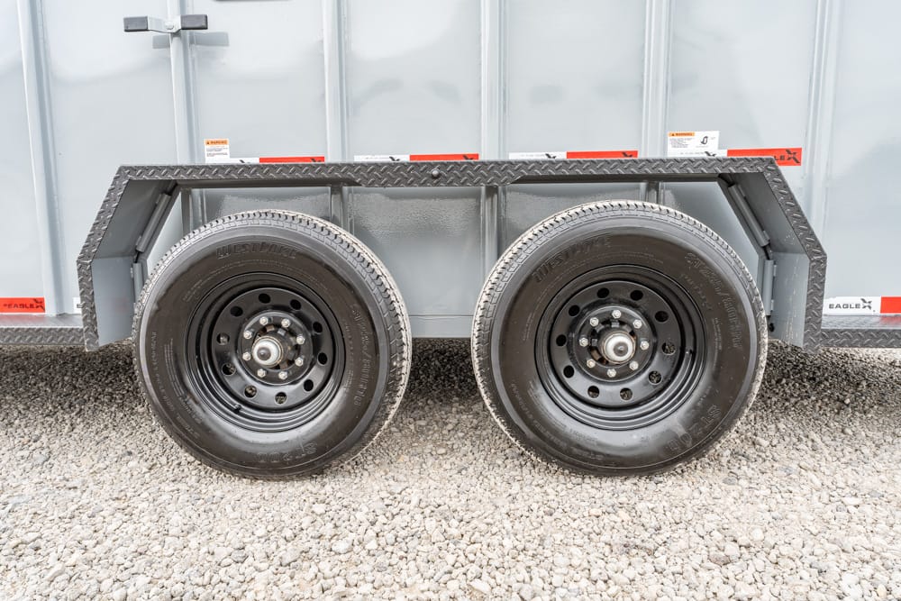 Dual-axle trailer wheels with black steel rims on a gray trailer, parked on gravel ground.