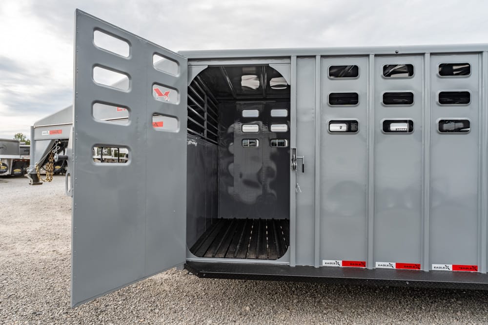 Open livestock trailer with its rear door swung wide, showing an empty gray metal interior on a gravel lot with ventilation slots along the side panels.