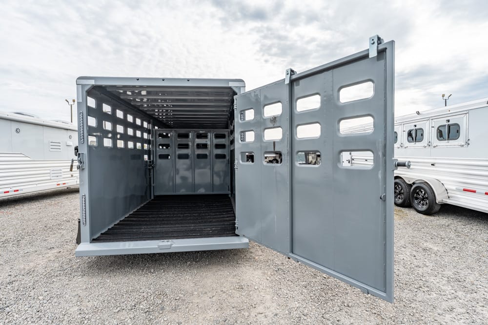 Grey livestock trailer with open rear doors, showing an empty interior and longitudinal partitions on a gravel lot.