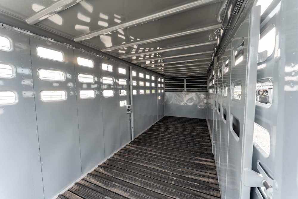 Interior of an empty gray horse trailer with a wooden plank floor and small rectangular windows along the sides, extending toward the back end.