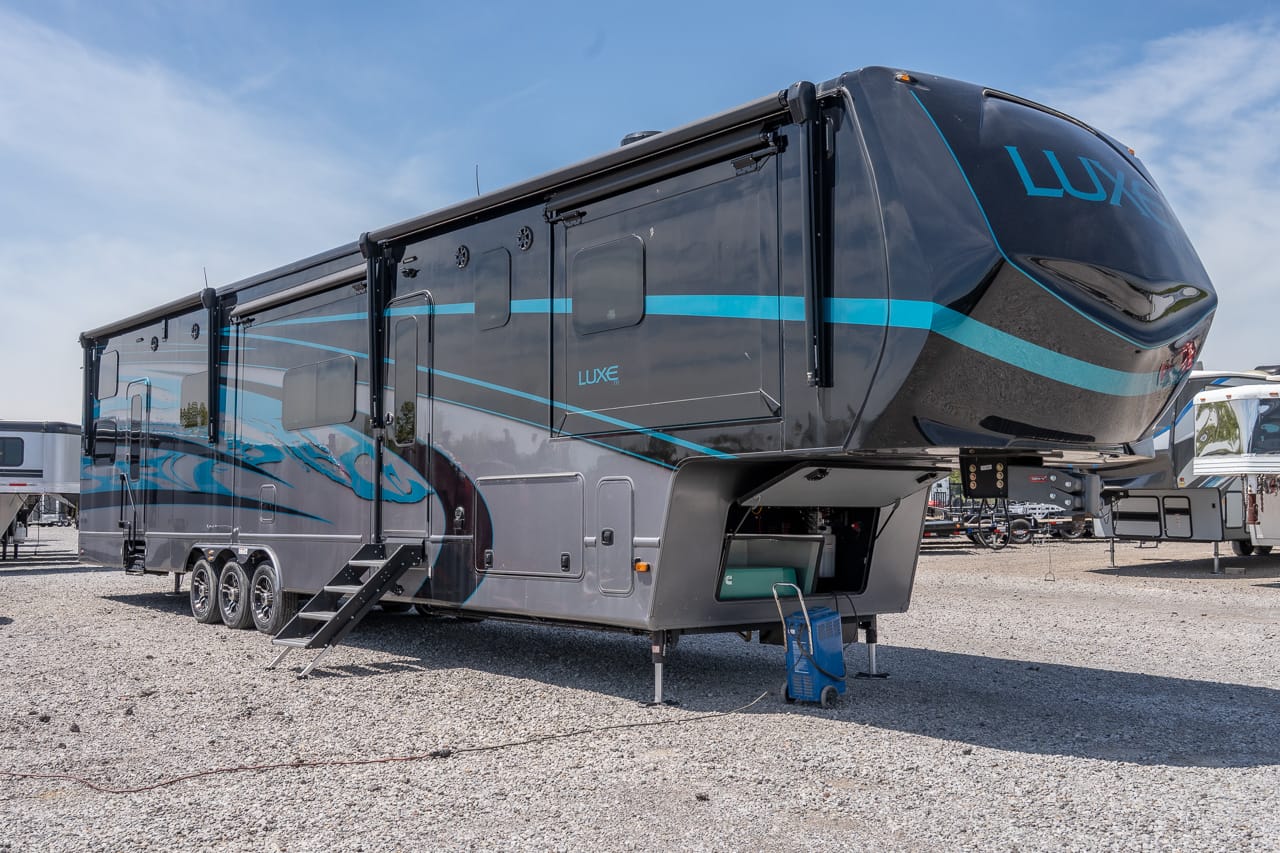 Luxurious dark gray fifth-wheel RV with blue accent lines, triple axles, and folding stairs on a gravel lot.