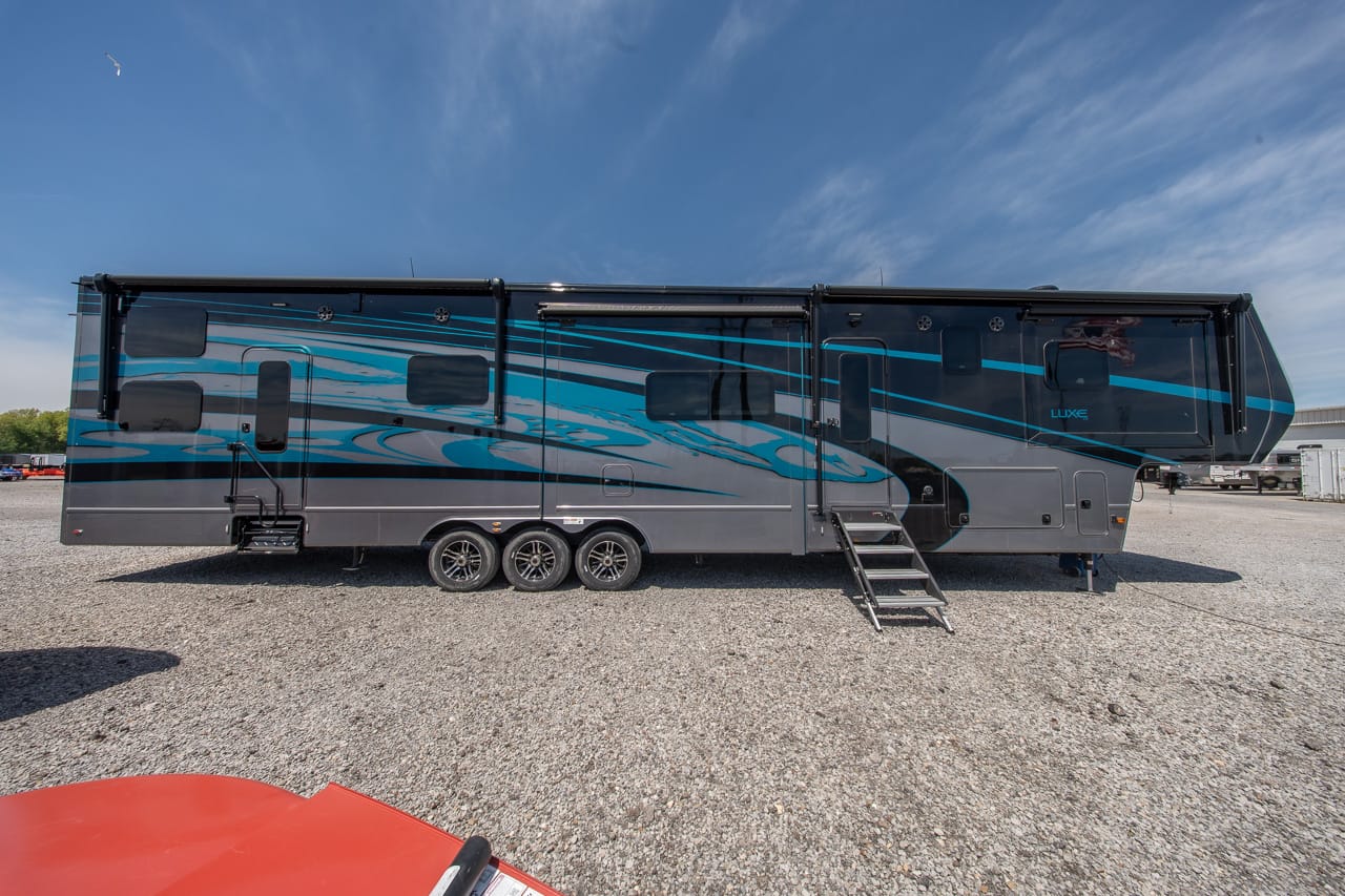 Long blue-and-gray luxury travel trailer parked on a gravel lot, with extended entry stairs and teal graphics along its side.