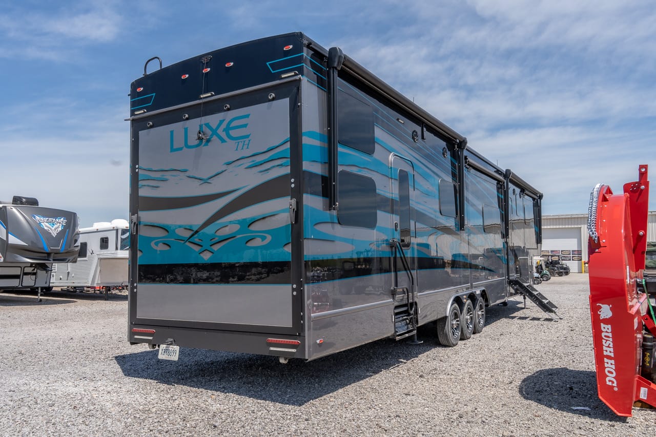 Large triple-axle Luxe TH luxury RV trailer parked on a gravel lot with blue graphics and multiple windows