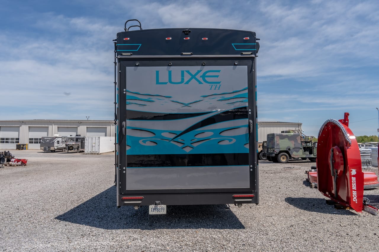 Rear view of a black Luxe TH trailer with blue and gray wave graphics, parked on a gravel lot with industrial buildings in the background.