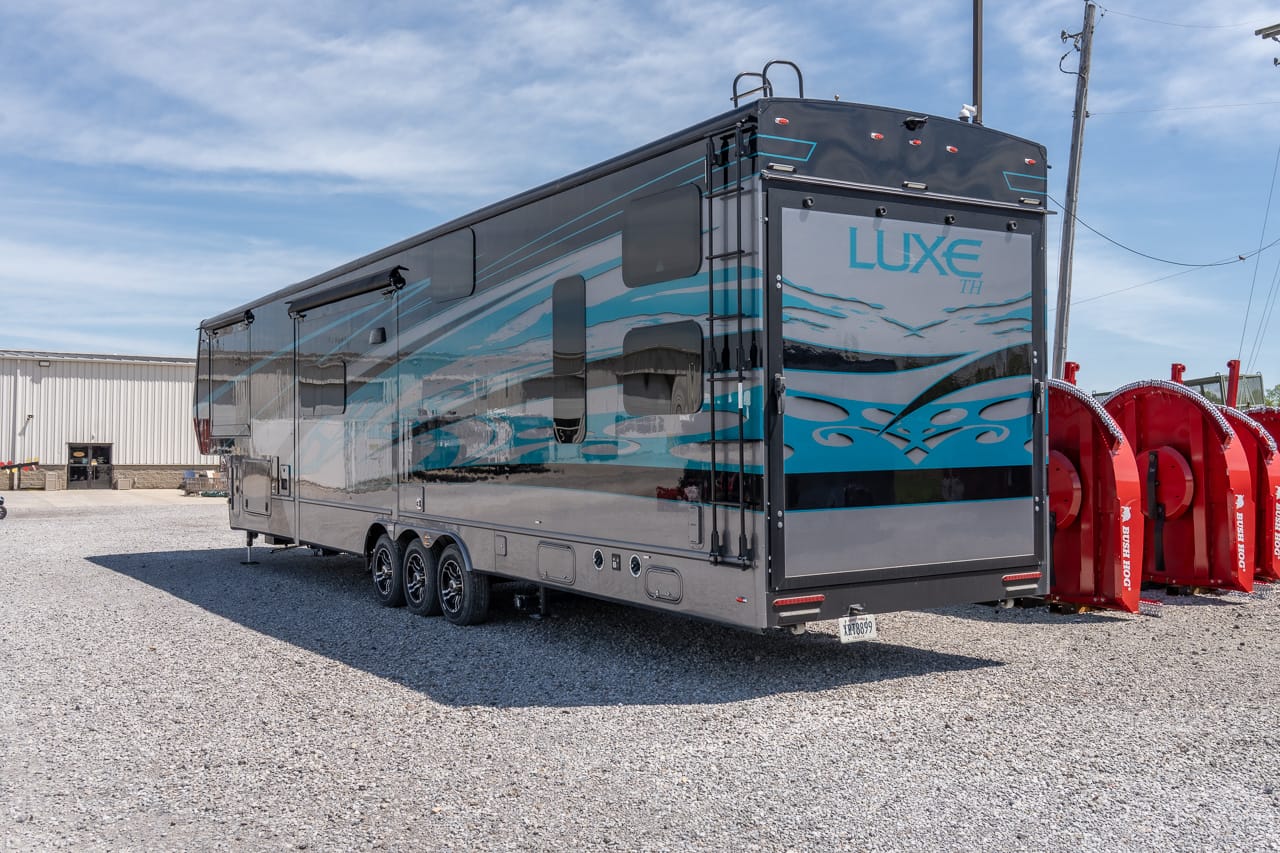 A large Luxe travel trailer with blue wave graphics parked on a gravel lot, viewed from the rear side.