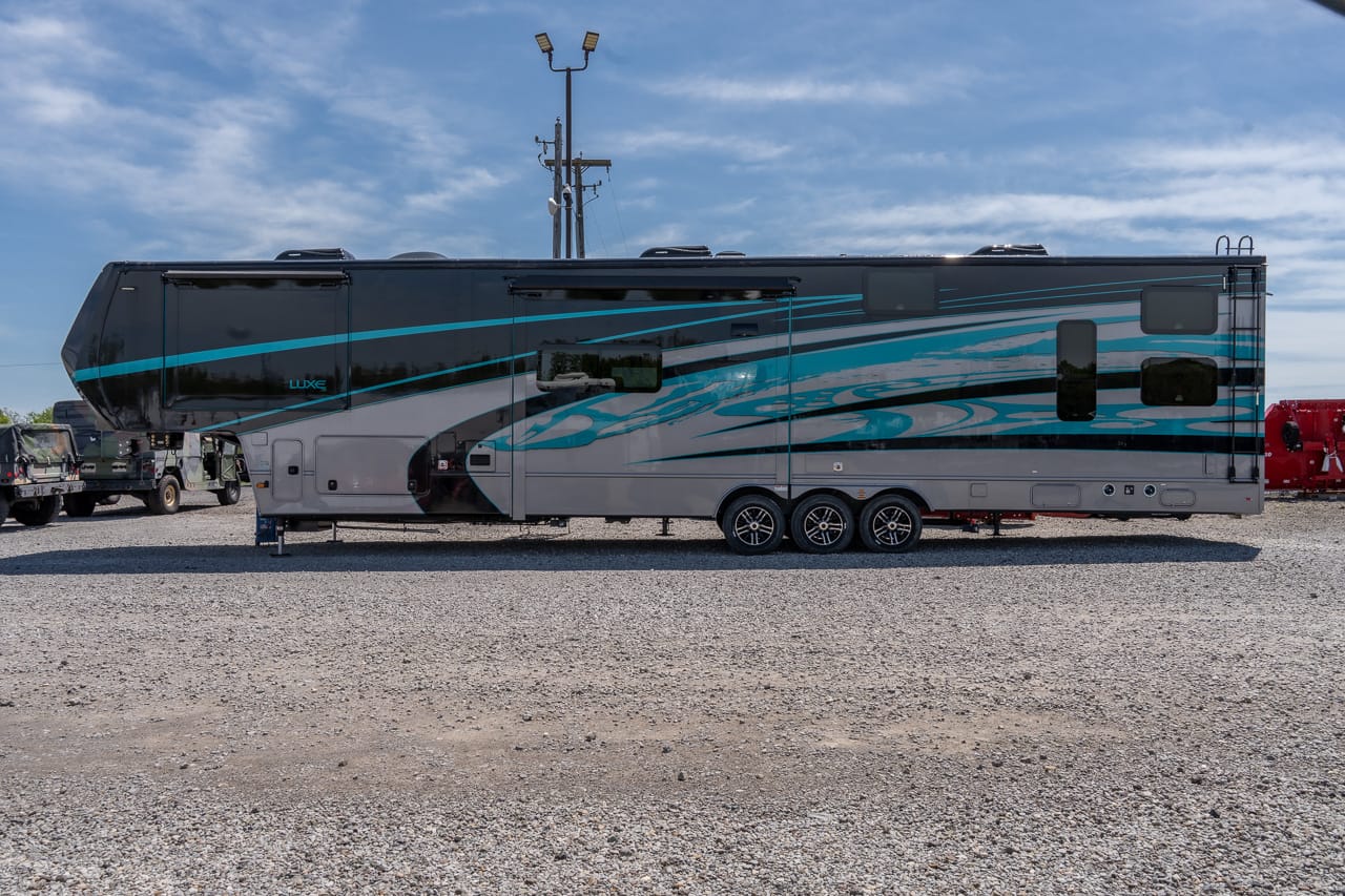 Long black and teal luxury fifth-wheel RV trailer parked on a gravel lot, side view with three axles and graphic swooshes.