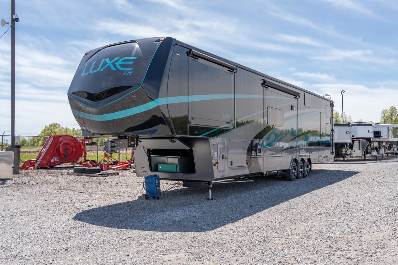 Large Luxe TH fifth-wheel trailer with teal graphics parked on a gravel lot under a blue sky