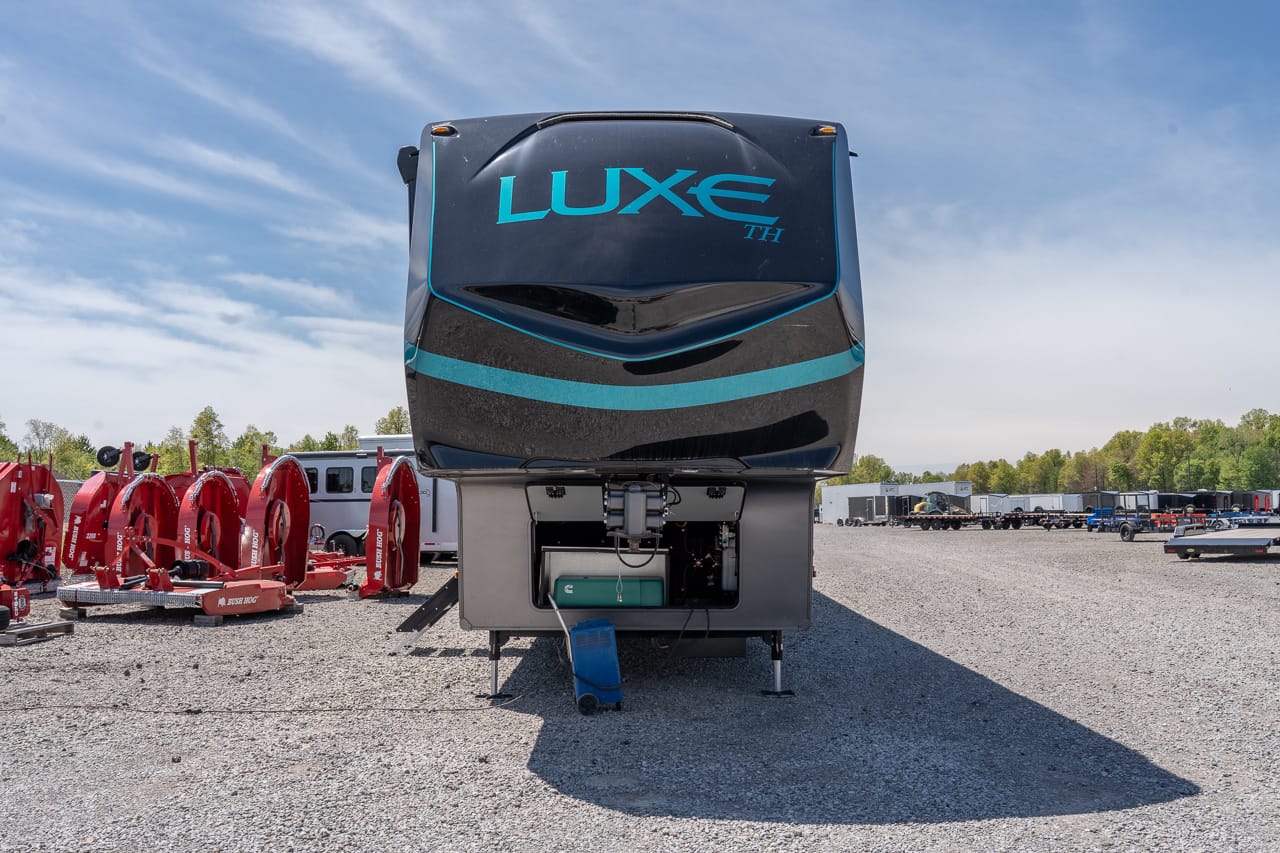 Front view of a dark blue Luxe TH fifth-wheel trailer parked on gravel with red industrial equipment to the left under a blue sky, large shadow in foreground.
