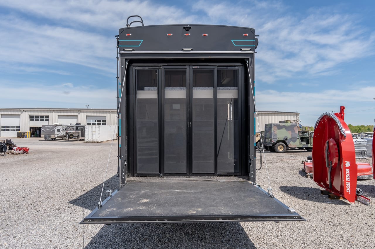 Open rear of a large black trailer with four vertical doors; rear ramp extended on a gravel lot, showing interior compartments behind doors on a sunny day.