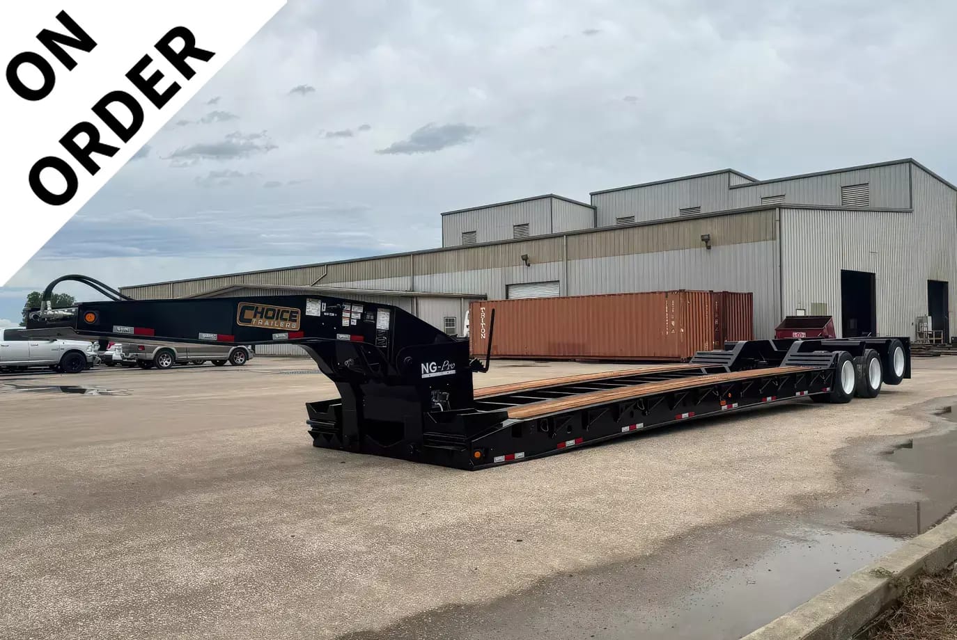 Long black lowboy trailer in a parking lot beside a large industrial warehouse; a diagonal white banner reads 'ON ORDER' across the top-left corner.