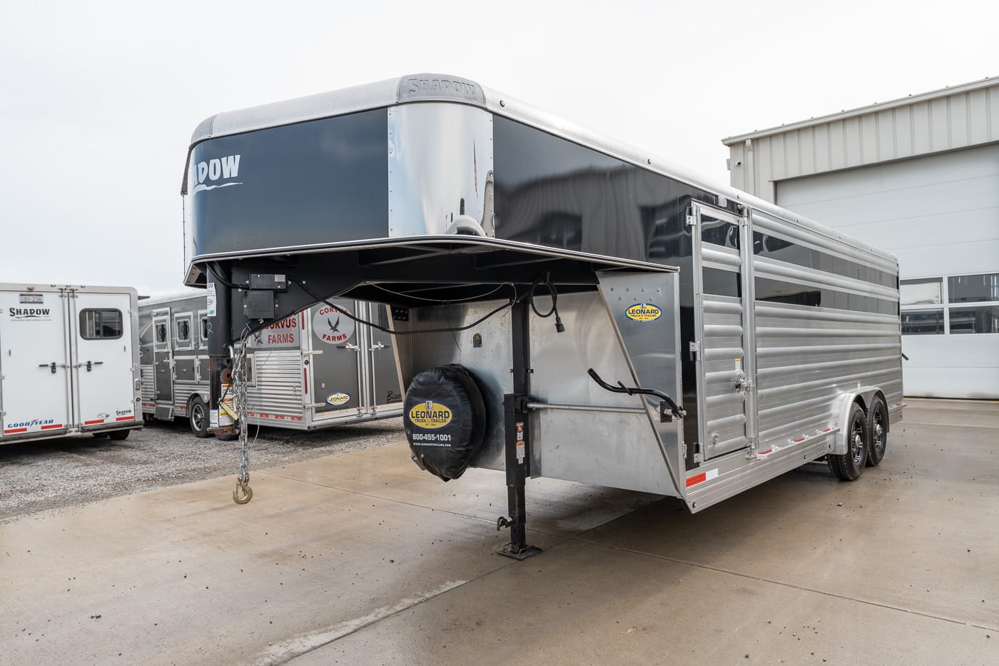 Silver gooseneck horse trailer with aluminum siding and a spare tire cover, parked in a lot with other trailers nearby.