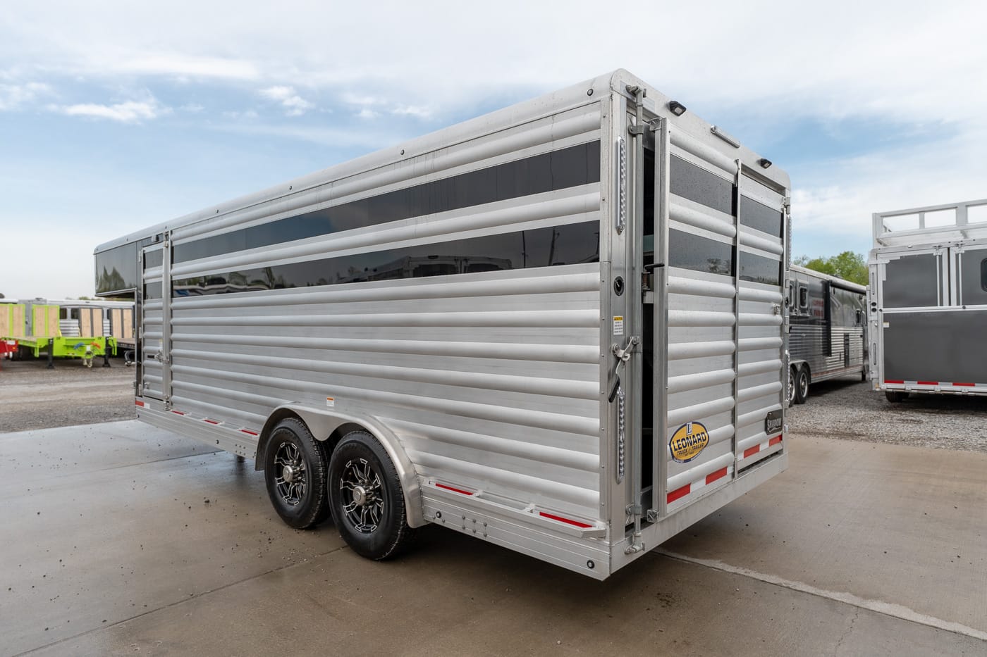 Two-axle aluminum horse trailer with ribbed sides and a rear double door, parked on a paved lot under a blue sky.