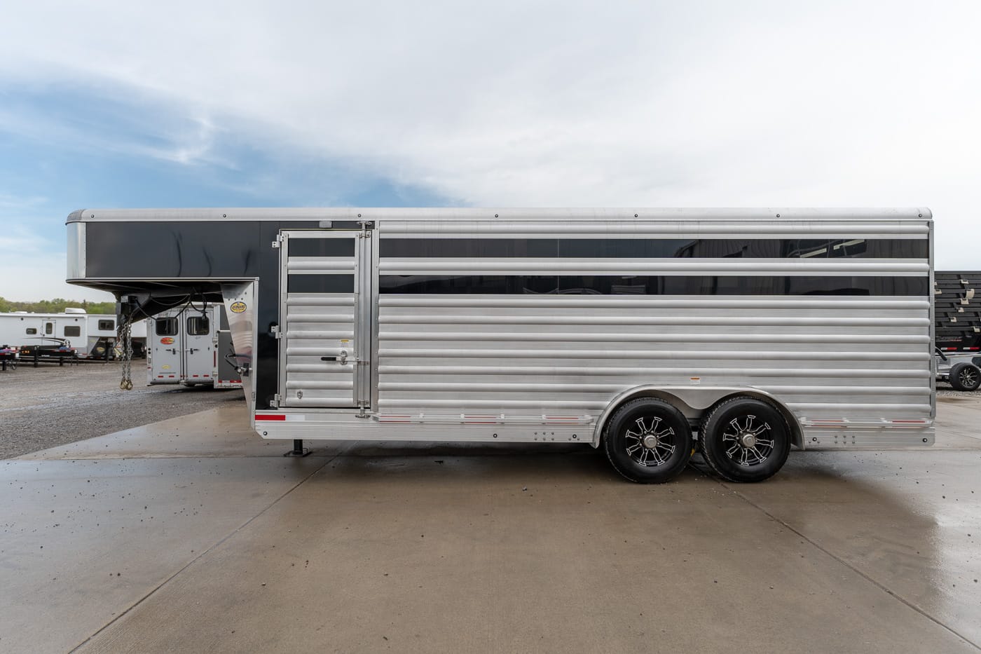 Silver aluminum horse trailer with two axles and a raised gooseneck, parked on a concrete lot with a side door open