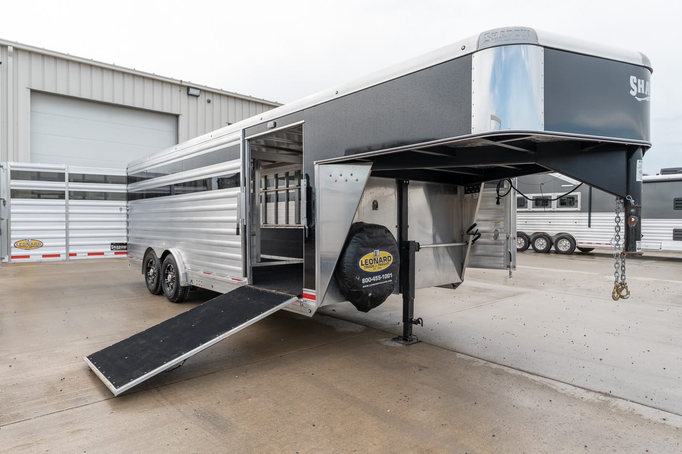 Silver and black horse trailer with open side door and lowered ramp for loading, parked on a concrete lot.