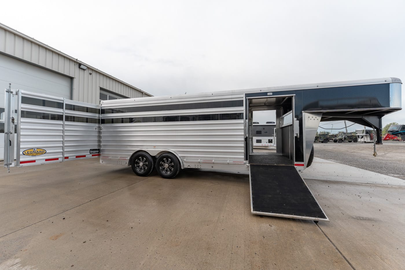 Two-axle aluminum livestock trailer with the rear ramp lowered, parked on a concrete lot beside a metal building.