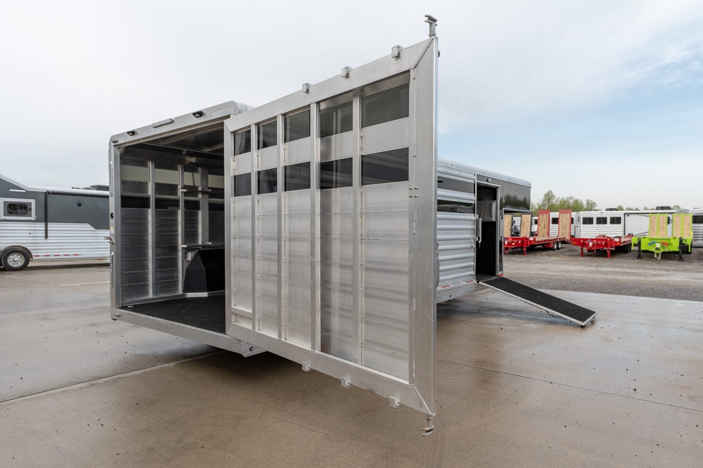Aluminum livestock trailer with its rear loading ramp lowered, exposing interior stalls on a paved lot with other trailers in the background.