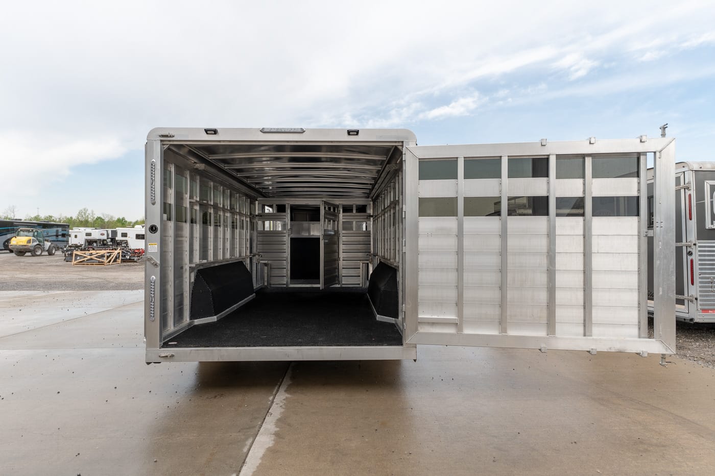 Rear view of an empty aluminum livestock trailer with divider gates inside, open on a paved lot with other trailers nearby.