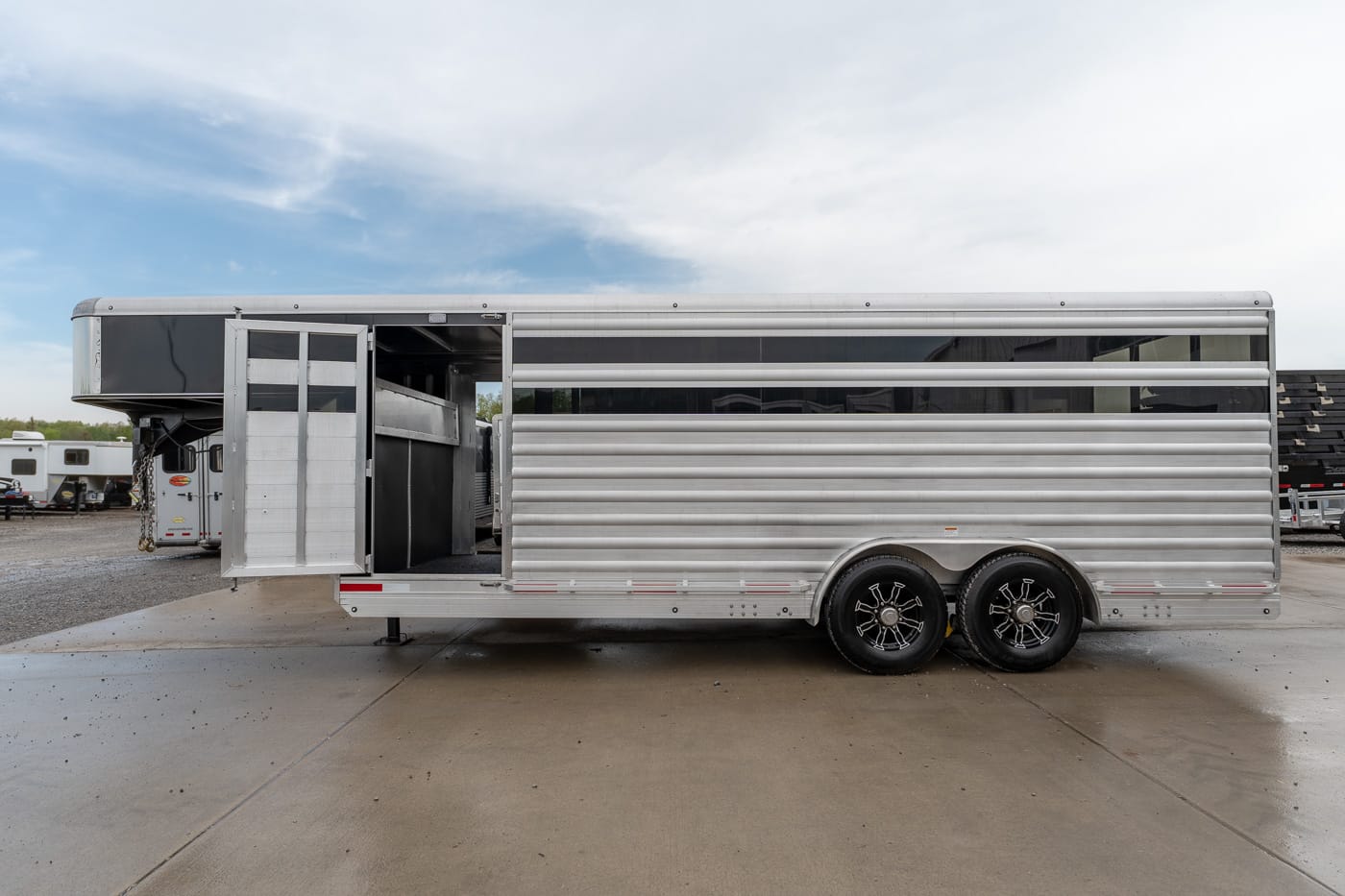Silver aluminum horse trailer with an open side door revealing interior stalls, parked on a wet concrete lot.