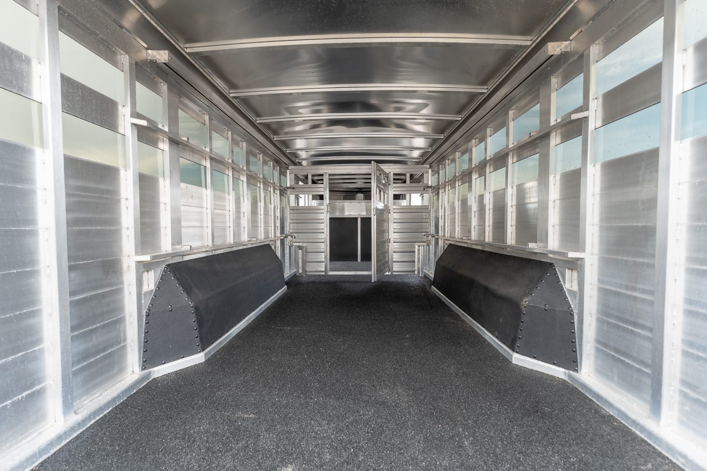 Empty aluminum horse trailer interior with metal dividers, side stalls, and a rubberized floor, viewed from the rear to the front