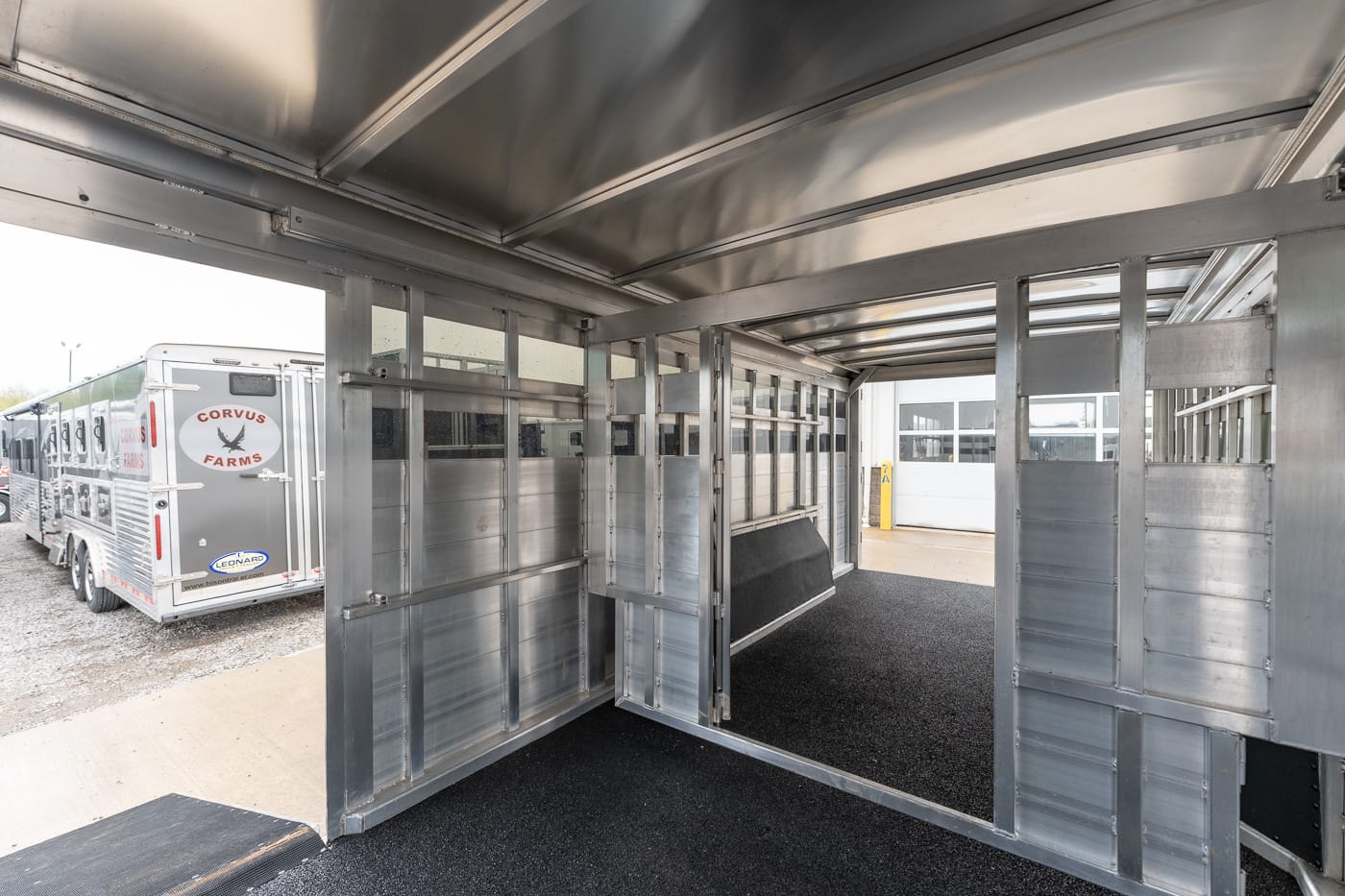 Interior of a silver horse trailer with partitioned stalls and black rubber flooring, ready for horses, at a staging area.