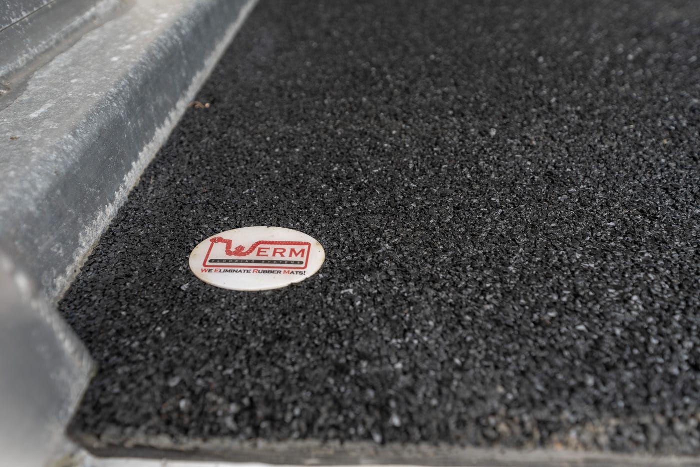 Close-up of a small circular white badge with a red logo lying on a textured black rubber mat near a metal edge.