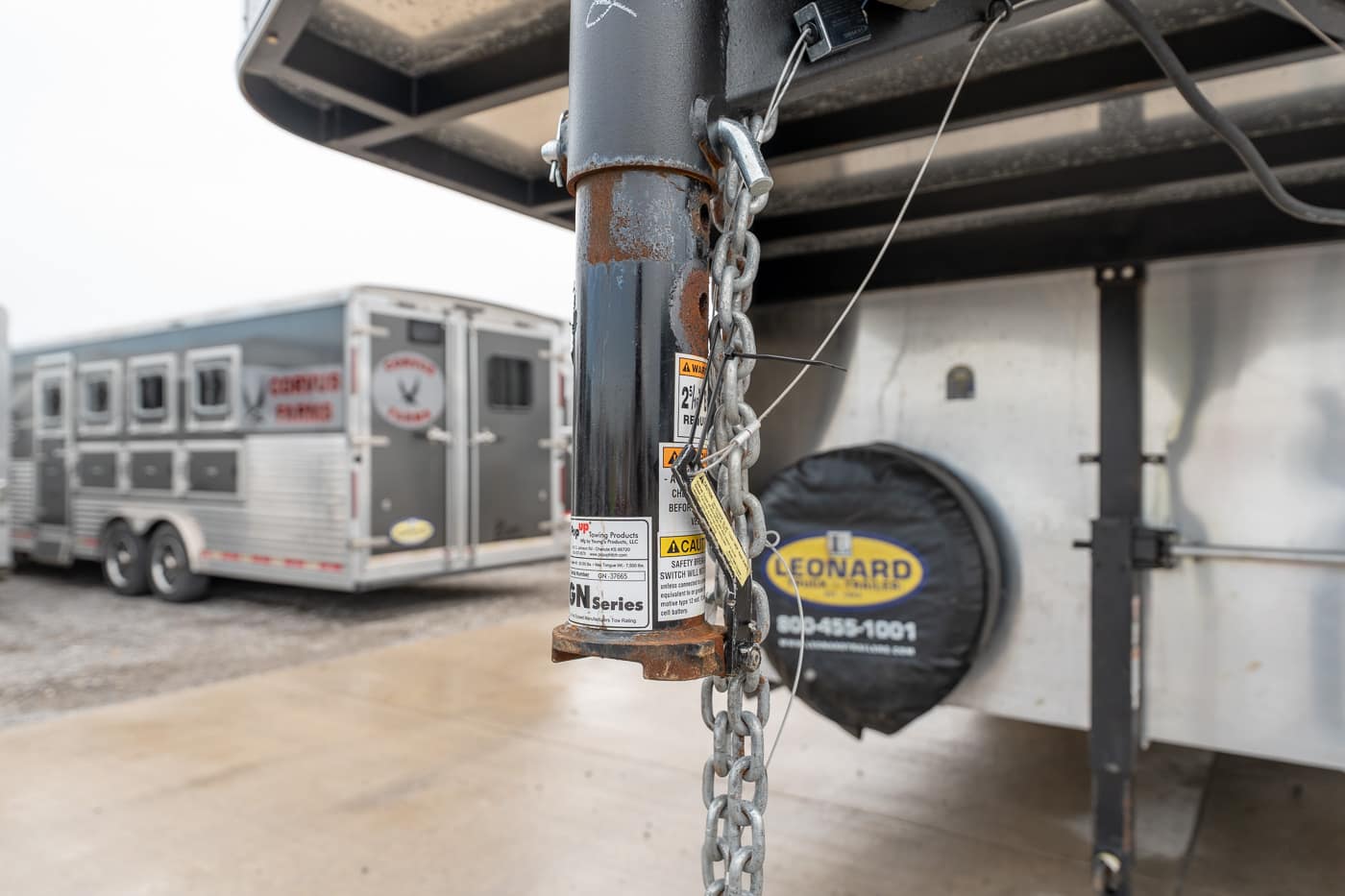 Close-up of a rusted trailer hitch post with safety chains and warning stickers, with a silver horse trailer in the background.