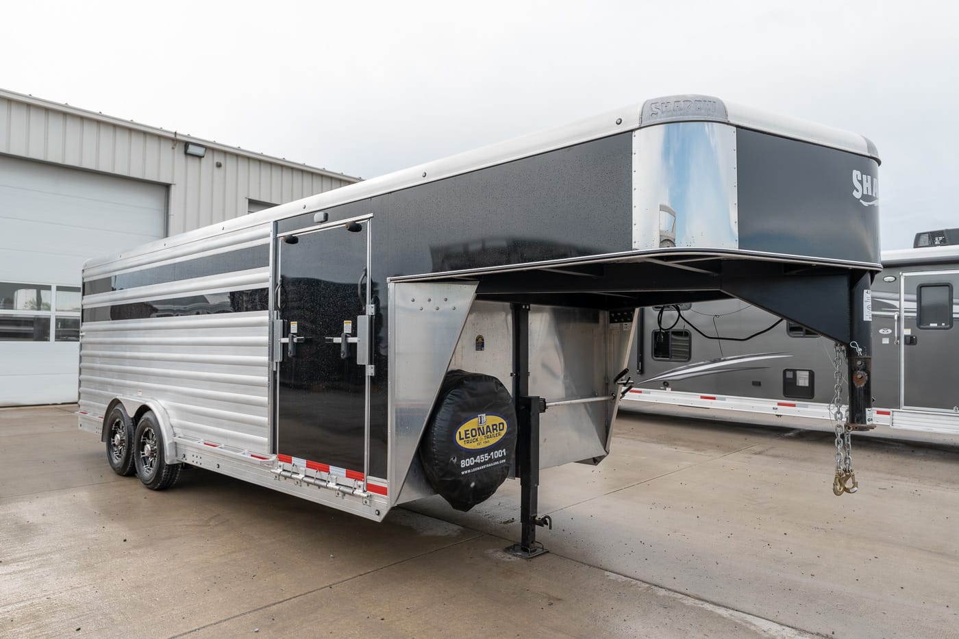 Enclosed gooseneck cargo trailer with twin axles and a spare tire cover, parked on a concrete lot by a metal building.
