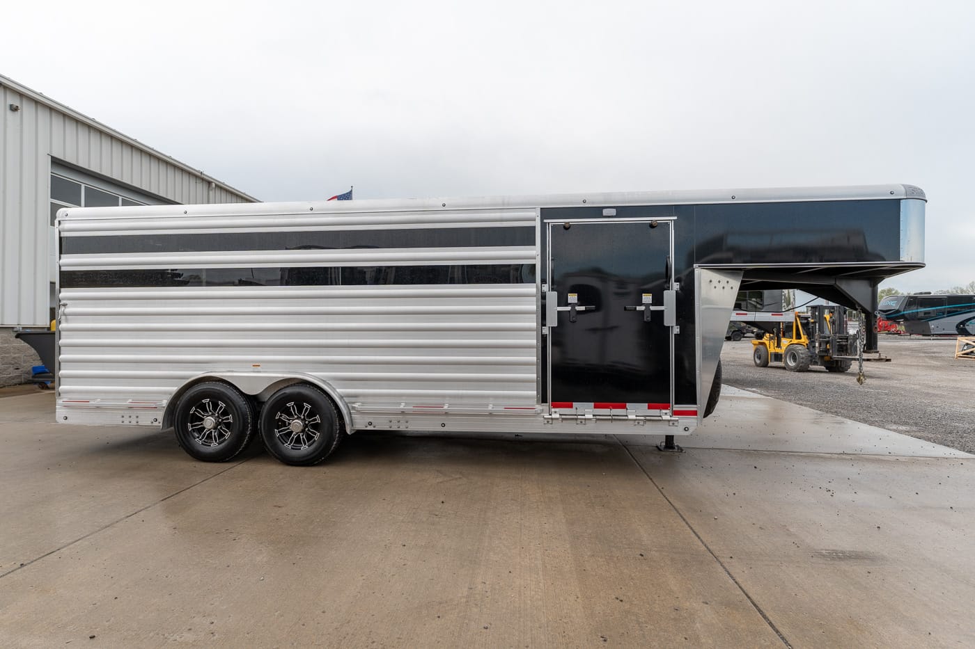 Silver aluminum livestock trailer with two tandem axles, black front section, parked beside a building.