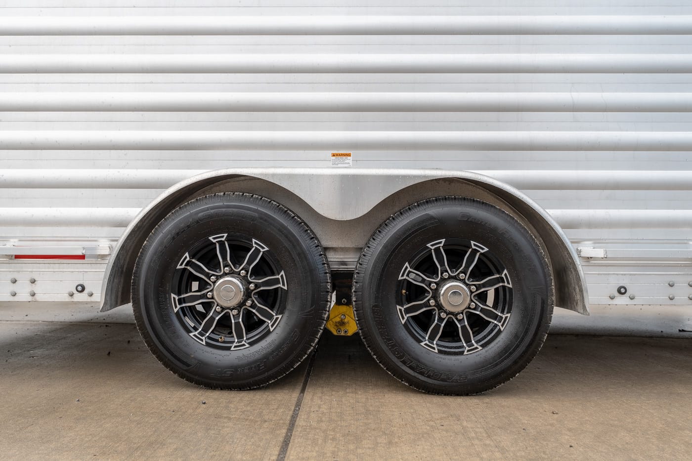 Dual-axle trailer with black alloy wheels and curved metal fenders resting on a concrete surface on a silver cargo trailer.