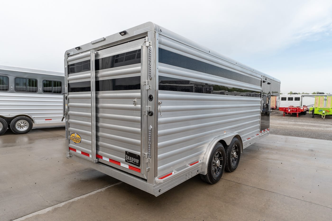 Rear three-quarter view of a silver aluminum livestock trailer with dual axles in a trailer yard, red/white reflective strips visible at the bumper.