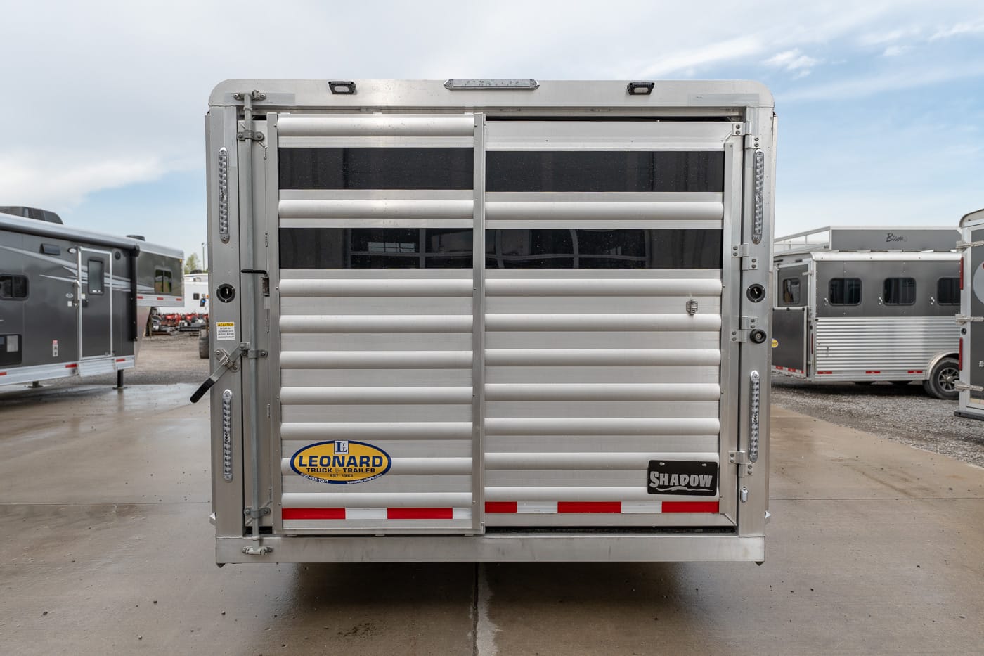 Rear view of a silver livestock/trailer with two vertical doors and horizontal vents; Leonard Truck & Trailer decal visible on left strap area and Shadow badge on right door.