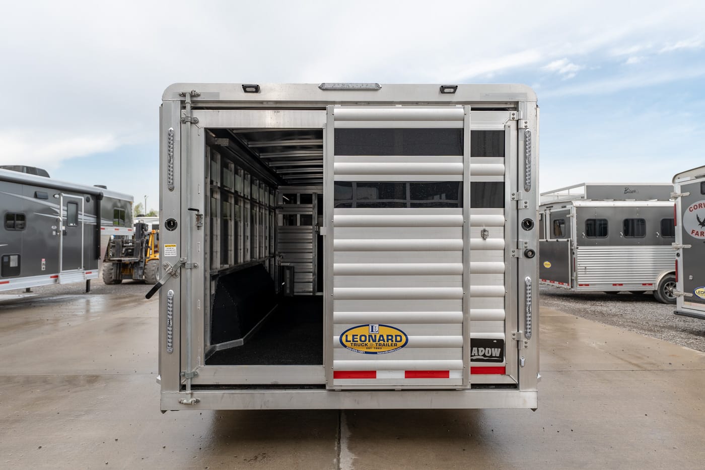 Rear view of a silver livestock trailer with the back doors open, showing empty stalls inside and a Leonard Truck Trailer logo on the door.