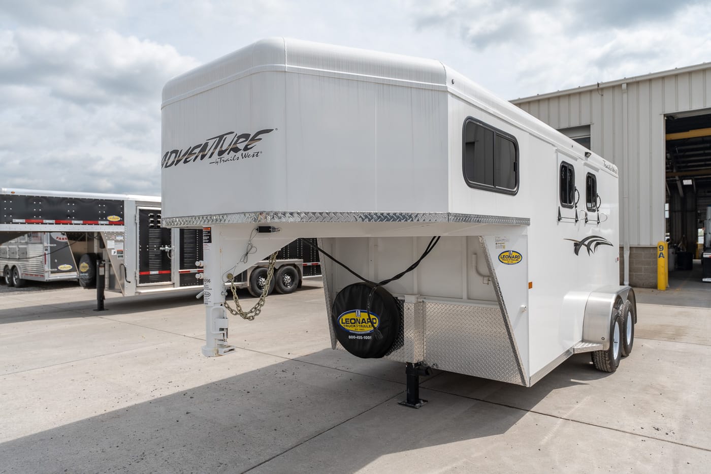 White gooseneck horse trailer with diamond-plate storage area, spare tire, and multiple windows, parked outside a building.