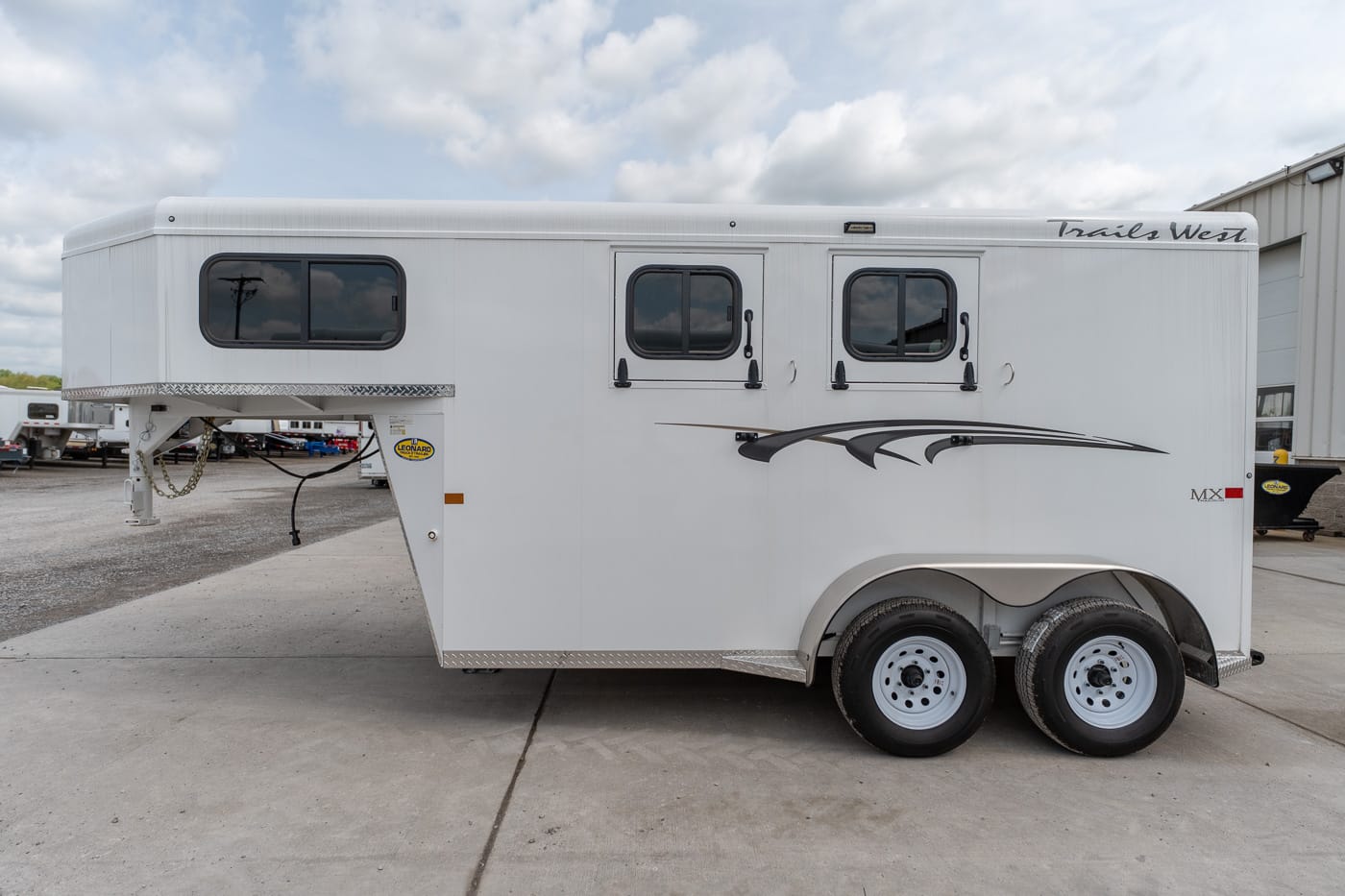 Side view of a white Trails West horse trailer with two axles, decorative graphics, and three windows on a concrete lot.