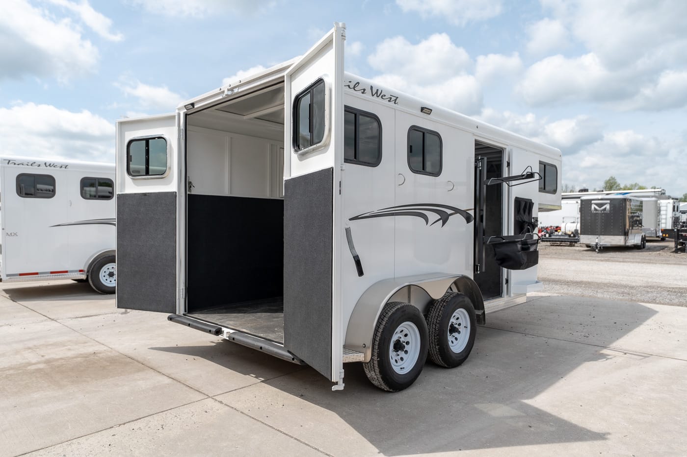 White two‑axle horse trailer with open rear doors showing a dark interior, parked in a lot with other trailers nearby.