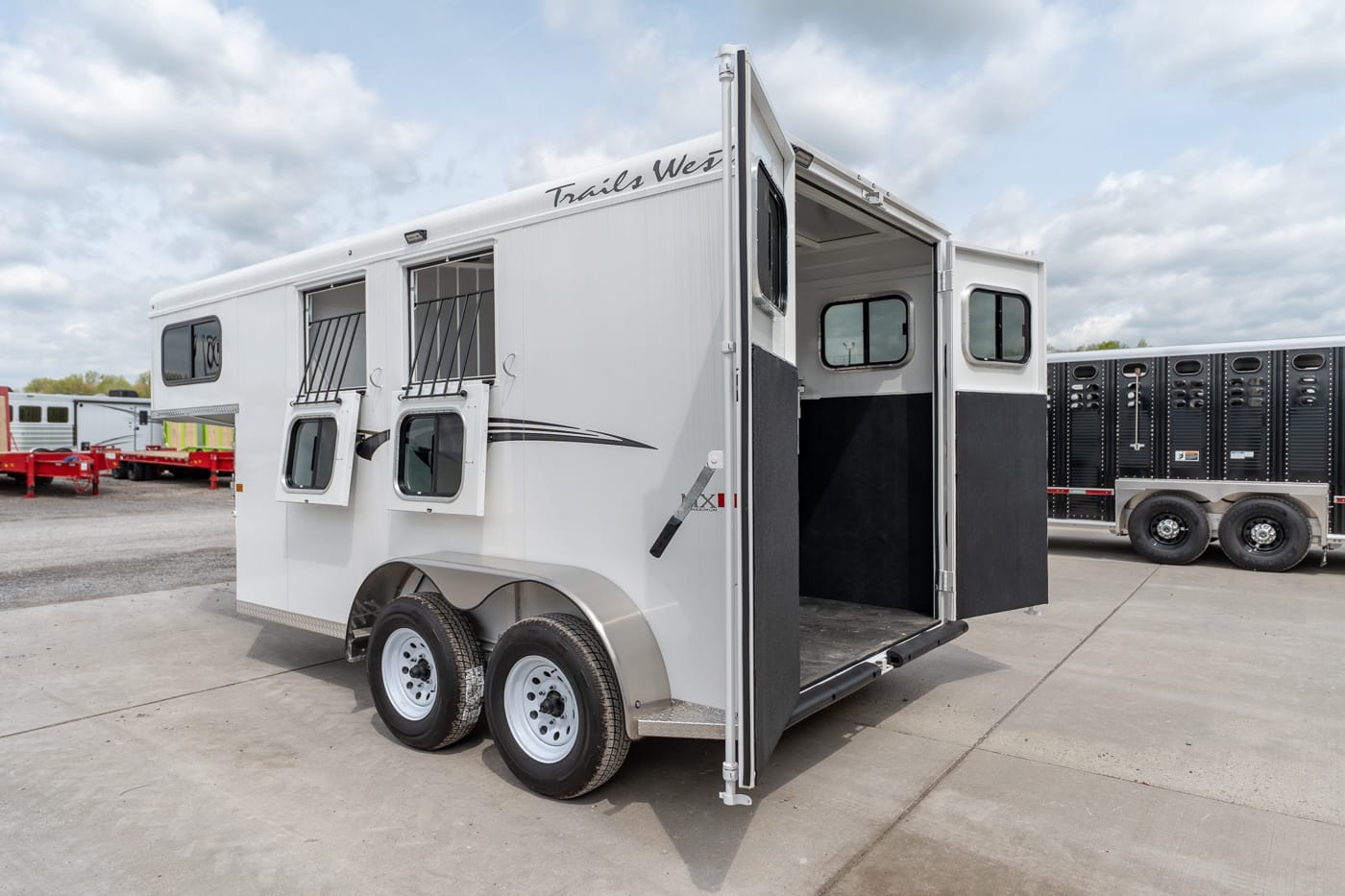 White double-axle horse trailer with open rear ramp, side windows with bars, and the Trails West logo above the door.