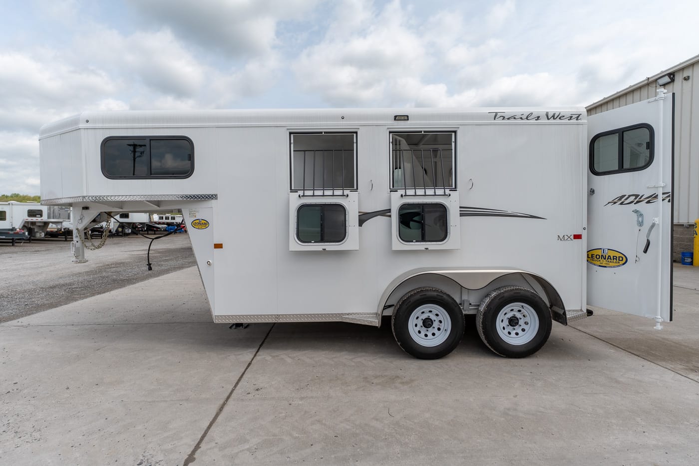 White two-axle horse trailer with barred stall doors and open windows, parked on a lot. It features a front hitch and dual wheels with metal trim along the bottom edge.