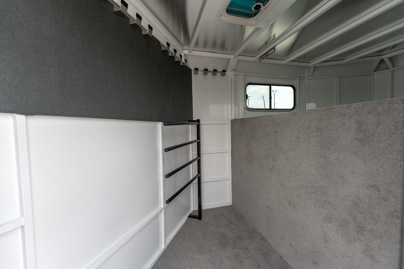 Interior of a light-gray trailer with white storage units along the left wall, a black metal bar rack, and a small window at the far end.