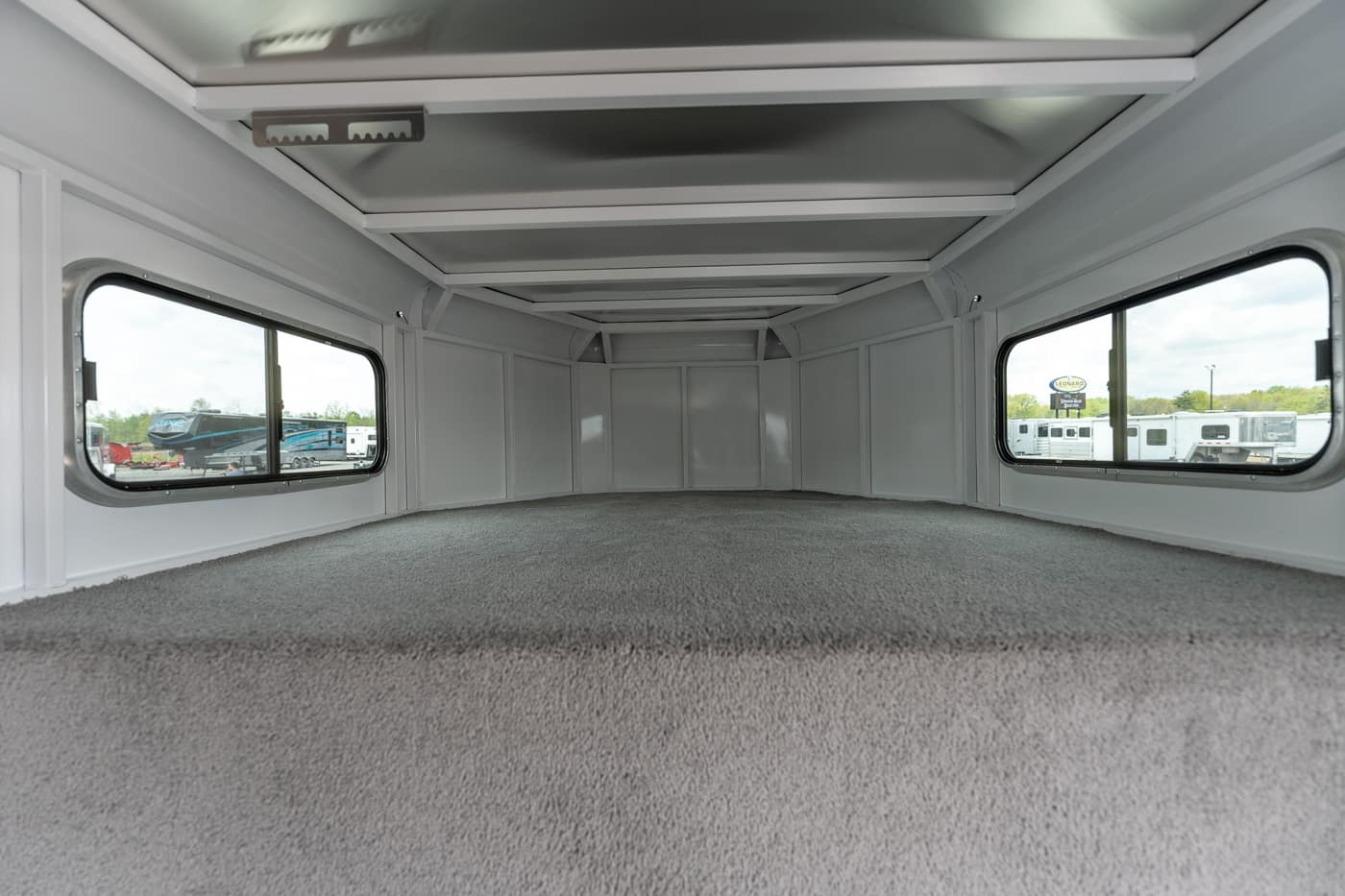 Interior of an empty camper bed/trailer cargo area with gray walls, carpeted floor, and side windows on both sides; overhead rails along the ceiling for storage.